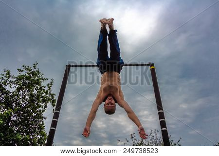 A Man With A Bare Torso In Blue Pants Upside Down Doing His Morning Exercises On A Horizontal Bar Ag