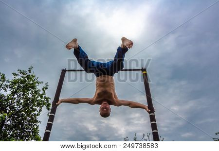 A Man With A Bare Torso In Blue Pants Upside Down Doing His Morning Exercises On A Horizontal Bar Ag