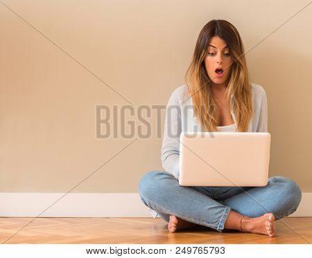 Young beautiful woman suprised sitting on the floor with computer looking at the laptop.