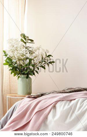 Photo With Bed With Pink Blanket In Blurred Foreground And Bedside Table With Flowers In Green Ceram