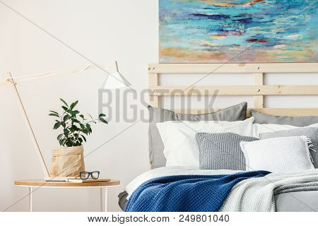 Plant And Lamp On Table Next To Bed With Blue Blanket In Bedroom Interior With Painting. Real Photo