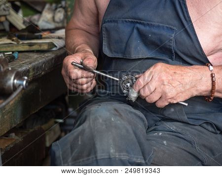 Older Man Has On Dark Navy Working Clothes Is Sitting At The Workbench In Workshop. Blurred Backgrou