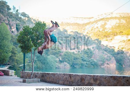 Young Man Doing A Side Flip Or Somersault While Practicing Parkour On A Lake In The Countryside.