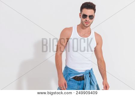 portrait of casual man wearing a white undershirt and a apir of sunglasses standing in spotlight on white background, in studio