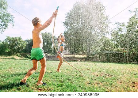 Two Childs Play Together In Garden, Hot Summer Day, They Make A Raim From Garden Watering Hose