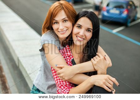 Two Atractive Young Women Hugging, Laughting And Sitting Near Road In The Street. One Brunette Girl 