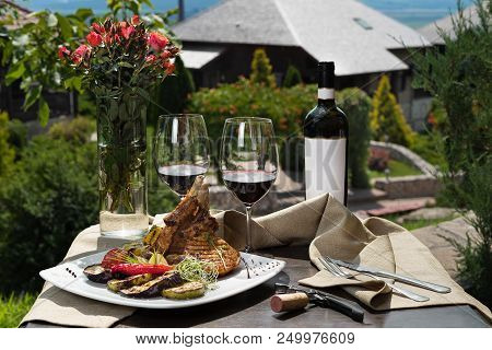 Romantic Dinner Outside. An Appetizing Dish Of Pork Ribs And Two Glasses Of Red Wine, Green Grass An
