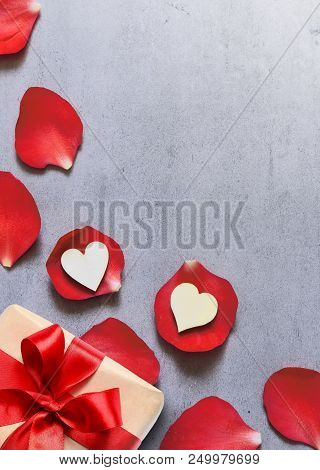 Gift In Craft Paper With Heart Decoration On Red Rose Petals Background On Grey Table. Romance Conce