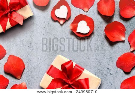 Gift In Craft Paper With Heart Decoration On Red Rose Petals Background On Grey Table. Romance Conce