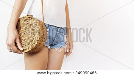 Young Girl In Denim Shorts And A White Sweatshirt With A Trendy Stylish Rattan Bag Against The Backd