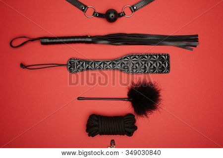 Top View Of Black Paddle, Gag, Rope, Feather Tickler And Flogging Whip Isolated On Red