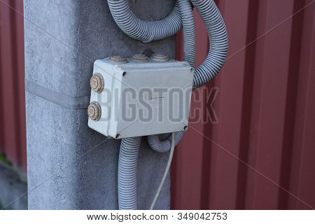 Closed White Metal Box And Plastic Hose On A Gray Concrete Pillar Against A Brown Wall On The Street