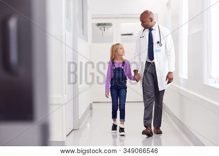 Male Paediatric Doctor Walking Along Hospital Corridor Holding Hands With Young Girl Patient