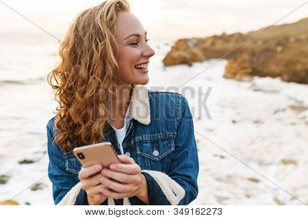 Image of young beautiful woman with blond curly hair using smartphone while walking by seaside