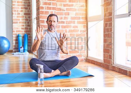 Middle age handsome sportman sitting on mat doing stretching yoga exercise at gym Moving away hands palms showing refusal and denial with afraid and disgusting expression. Stop and forbidden.