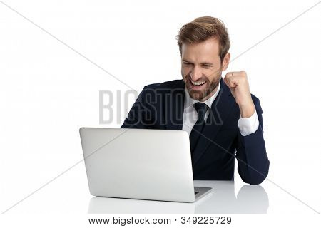excited businessman in navy blue suit reading emails and holding fist in the air, celebrating victory and sitting isolated on white background, full body
