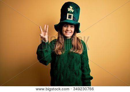 Young beautiful brunette woman wearing green hat on st patricks day celebration showing and pointing up with fingers number three while smiling confident and happy.
