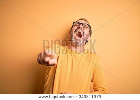 Middle age hoary man wearing casual sweater and glasses over isolated yellow background laughing at you, pointing finger to the camera with hand over body, shame expression