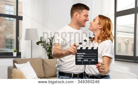 production, filmmaking and entertainment concept - portrait of happy couple in white t-shirts with clapperboard over home background