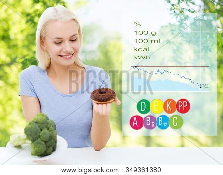 eating, diet and people concept - happy smiling woman choosing between broccoli and donut with calories, vitamins and food nutritional value over green natural background