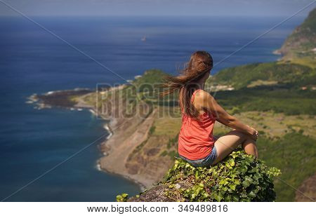 Young Woman With Long Dark Hair In Red Shirt And Denim Jeans Sitting On A Precipice. View From Behin