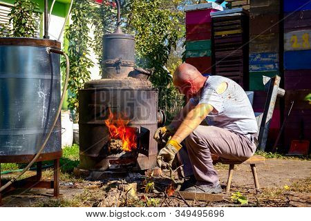 Man Throws Dry Branches Into The Firebox Of A Homemade Distillery Making Moonshine Schnapps, Alcohol