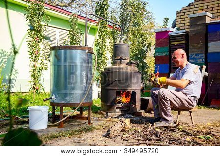 Man Throws Dry Branches Into The Firebox Of A Homemade Distillery Making Moonshine Schnapps, Alcohol