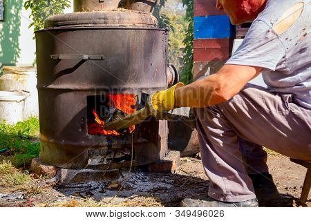 Man Throws Dry Branches Into The Firebox Of A Homemade Distillery Making Moonshine Schnapps, Alcohol
