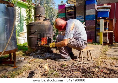 Man Throws Dry Branches Into The Firebox Of A Homemade Distillery Making Moonshine Schnapps, Alcohol