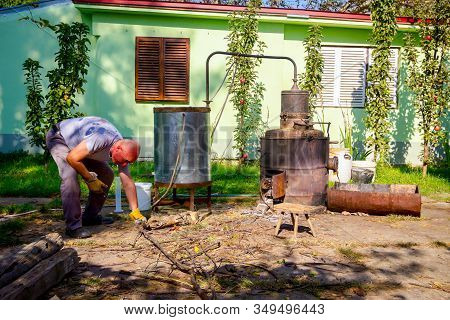 Man Throws Dry Branches Into The Firebox Of A Homemade Distillery Making Moonshine Schnapps, Alcohol