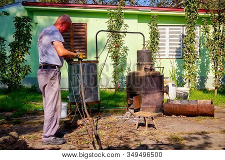 Man Throws Dry Branches Into The Firebox Of A Homemade Distillery Making Moonshine Schnapps, Alcohol