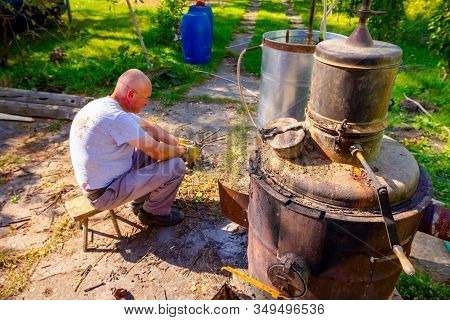 Man Throws Dry Branches Into The Firebox Of A Homemade Distillery Making Moonshine Schnapps, Alcohol