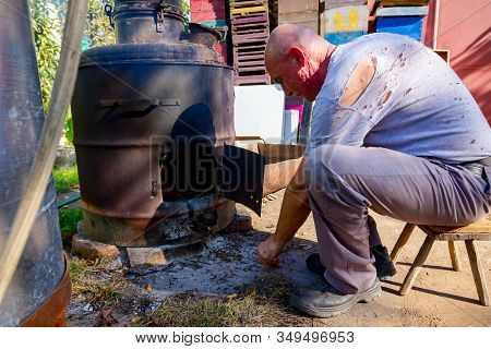 Man Throws Dry Branches Into The Firebox Of A Homemade Distillery Making Moonshine Schnapps, Alcohol