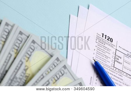 1120 Tax Form Lies Near Hundred Dollar Bills And Blue Pen On A Light Blue Background. Us Corporation