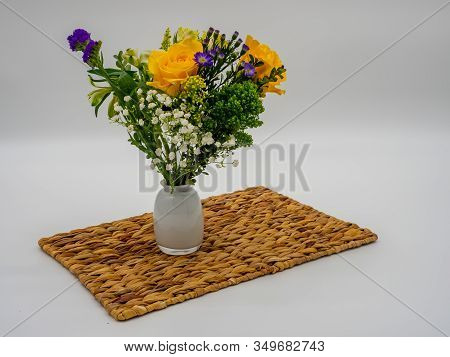 Purple, Yellow And Green Floral Arrangement In A White Glass Vase Sitting On A Brown Whicker Placema