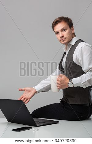 Businessman In A White T-shirt And Grey Vest Drinking Coffee Sitting On A White Table With Laptop An