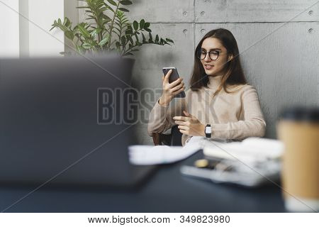 Young Female Executive Sitting At Workplace At Lunch Time And Listening To Music On Smartphone Using