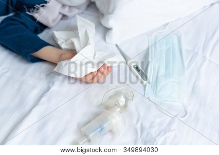 Hand With Handkerchief In Hand And Protective Mask, Thermometer And Nebuliser On The Bed. Virus, Con