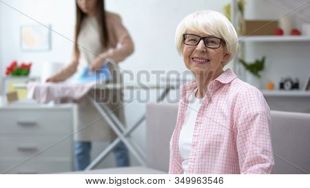 Smiling Old Woman Looking At Camera, Granddaughter Ironing Clothes On Background