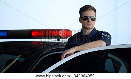 Serious Patrolman In Sunglasses Monitoring Road Standing Near Door Of Police Car