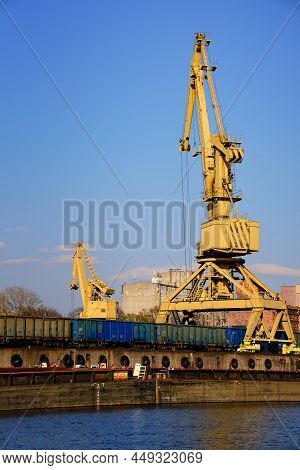 River Port Crane Loading Open-top Gondola Cars On Sunny Day. Empty River Drag Boats Or Barges Moored