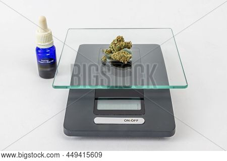One Gram Of Dried Medical Marijuana Buds On A Scale, 10ml Blue Glass Bottle Of Cbd Oil On A White St