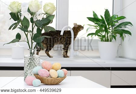 A Wicker Basket With Multi-colored Easter Eggs And A Vase With White Roses Against The Background Of