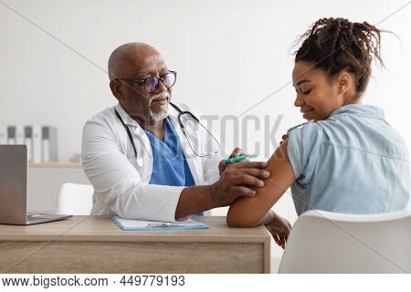 Black Lady Receiving Coronavirus Vaccine Injection Sitting With Doctor Indoor