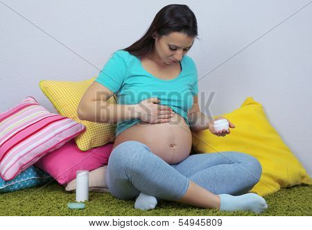 Young regnant woman sitting on floor and applying cream on her belly on wall background