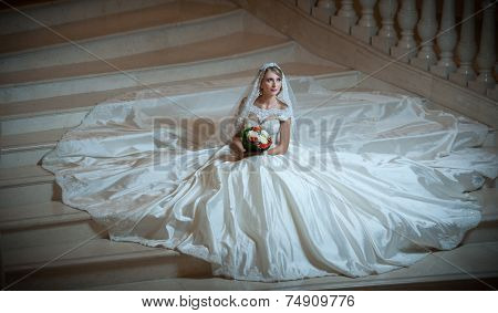 Young beautiful luxurious woman in wedding dress sitting on stair steps in semi-darkness. Bride