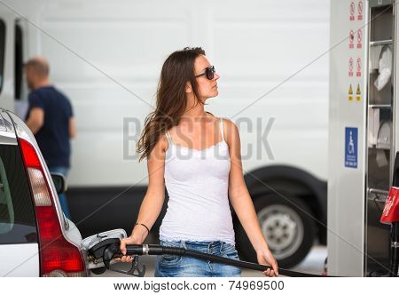 Attractive, young woman refueling her car in a gas station, checking the amount of gas, disliking the price tag and the gas mileage of her engine