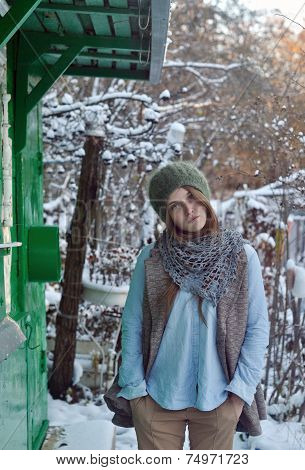winter portrait of beautiful sad girl wearing green hat and coat standing near green house