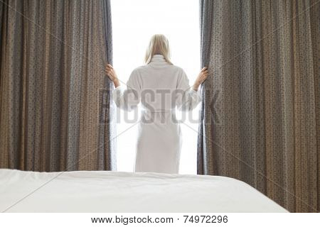 Rear view of young woman in bathrobe opening window curtains at hotel room