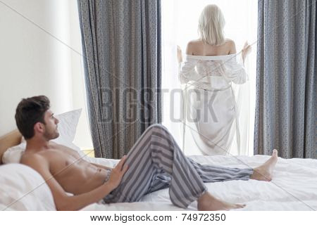 Full length of shirtless young man looking at woman standing by hotel window